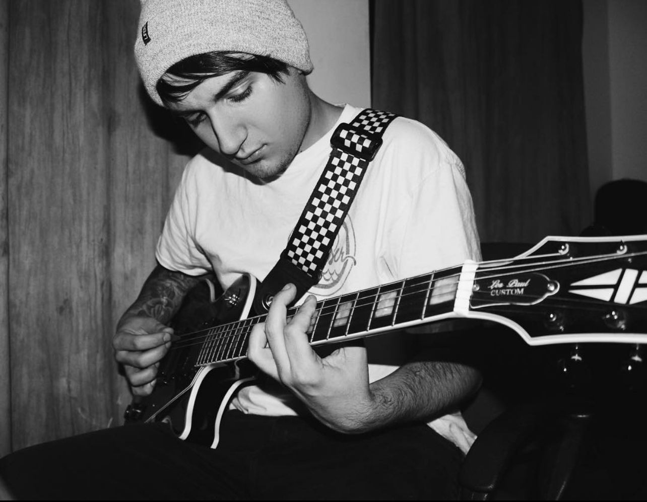 Person playing electric guitar while wearing a beanie and patterned strap in a dimly lit room.
